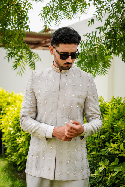 Light sage/grey prince coat with vertical embroidery and mirror work all over. Band collar with subtle gold detailing. Paired with matching kurta pajama
